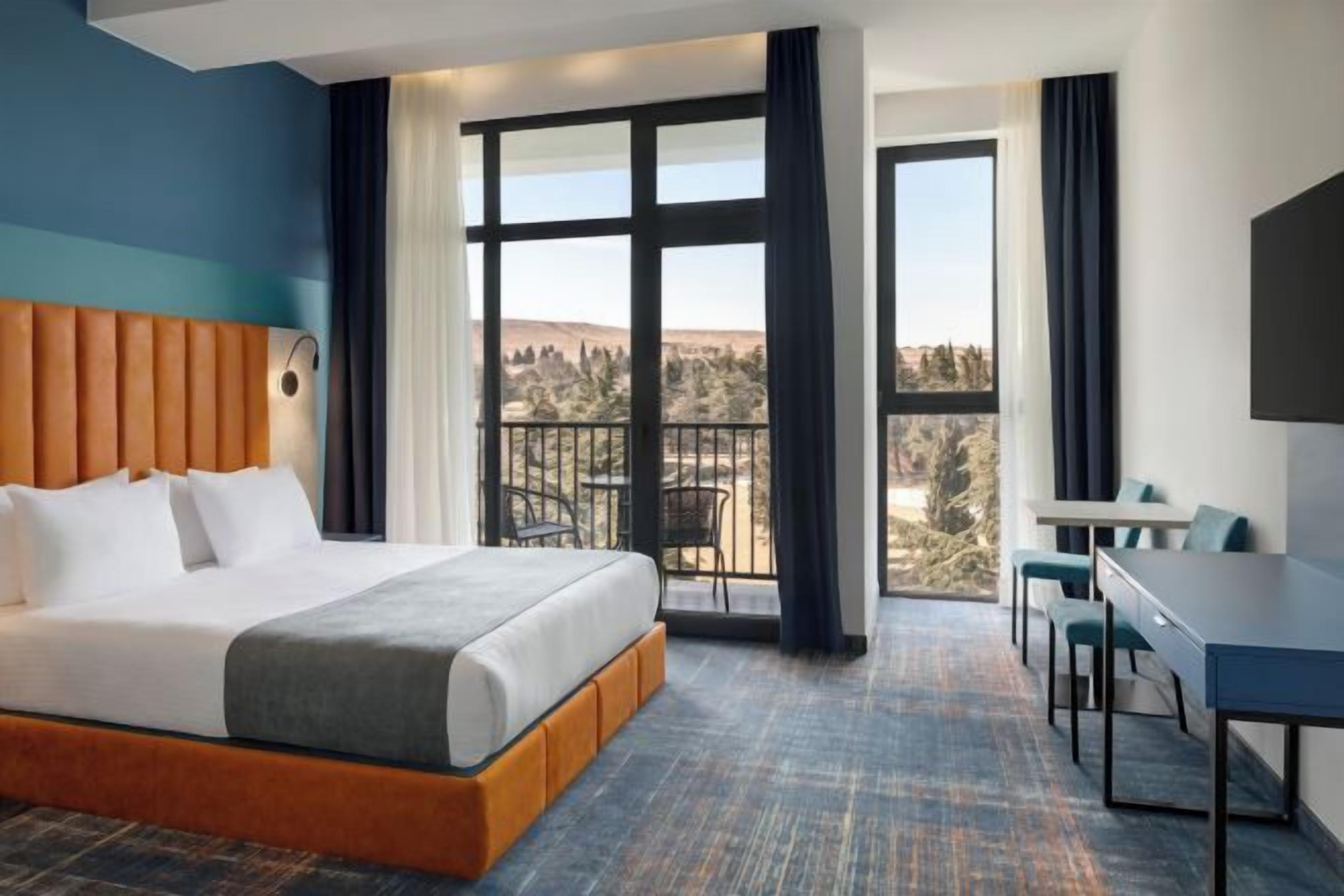 Habitación de hotel con una cama grande, un balcón con vistas, un escritorio y una silla.