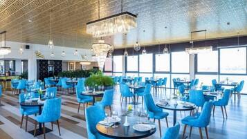 Un comedor espacioso con mesas redondas y sillas azules, ventanas amplias y un techo alto con diseño decorativo.