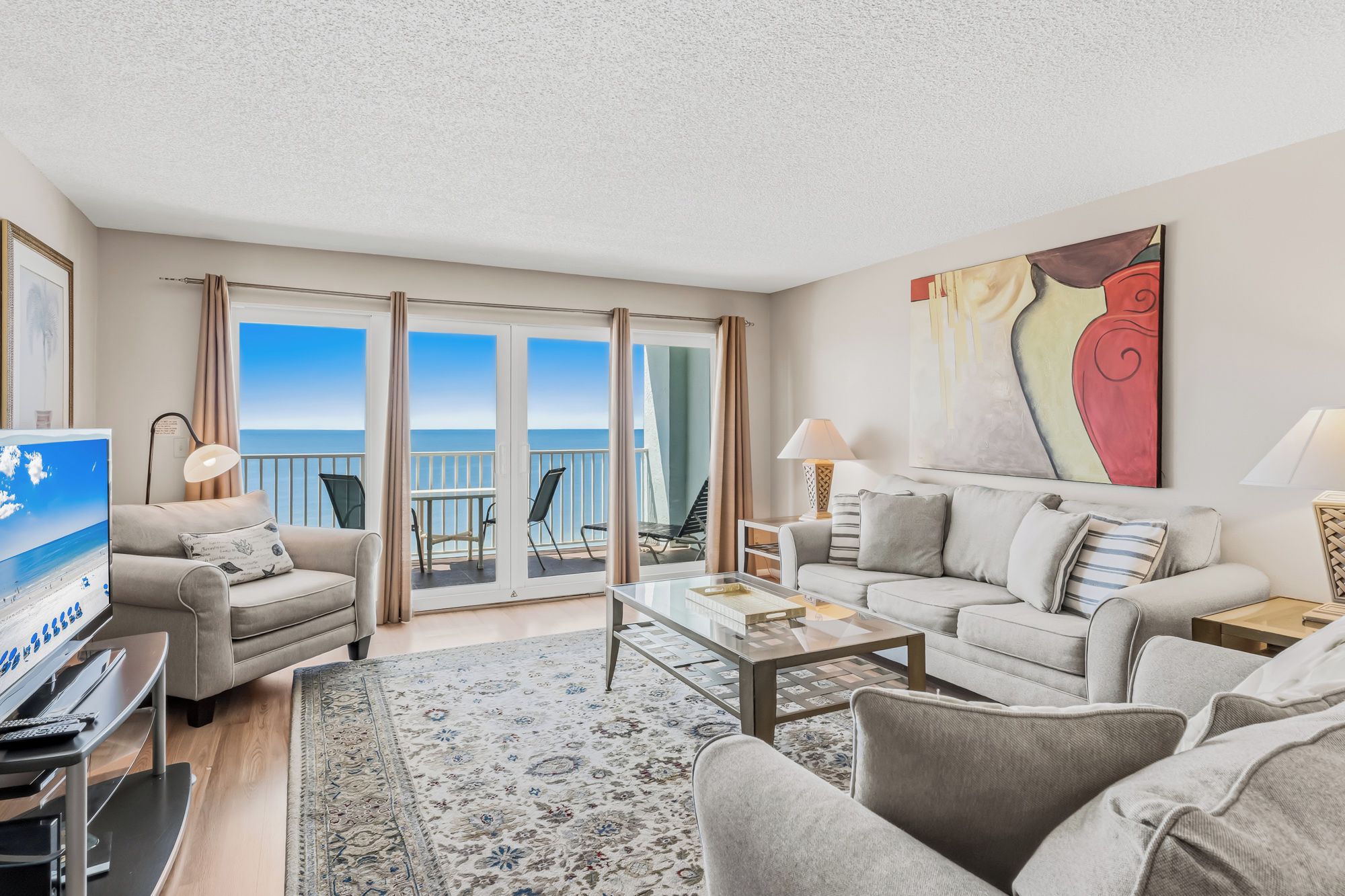 A modern hotel room with a balcony, a flat-screen TV, a sofa, a coffee table, and a large abstract painting.
