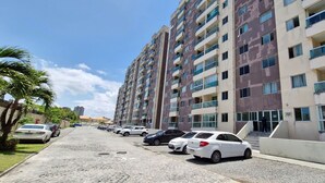A multi-story residential building with balconies, a parking area with cars, and a clear sky. - COJOC0100 - Cond Torres do Atlântico (Lauro de Freitas)
