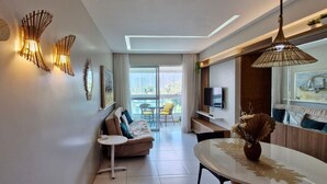 A modern living room with a sofa, a dining table, and a TV. There is a balcony with a view of buildings. - COJOC0100 - Cond Torres do Atlântico (Lauro de Freitas)