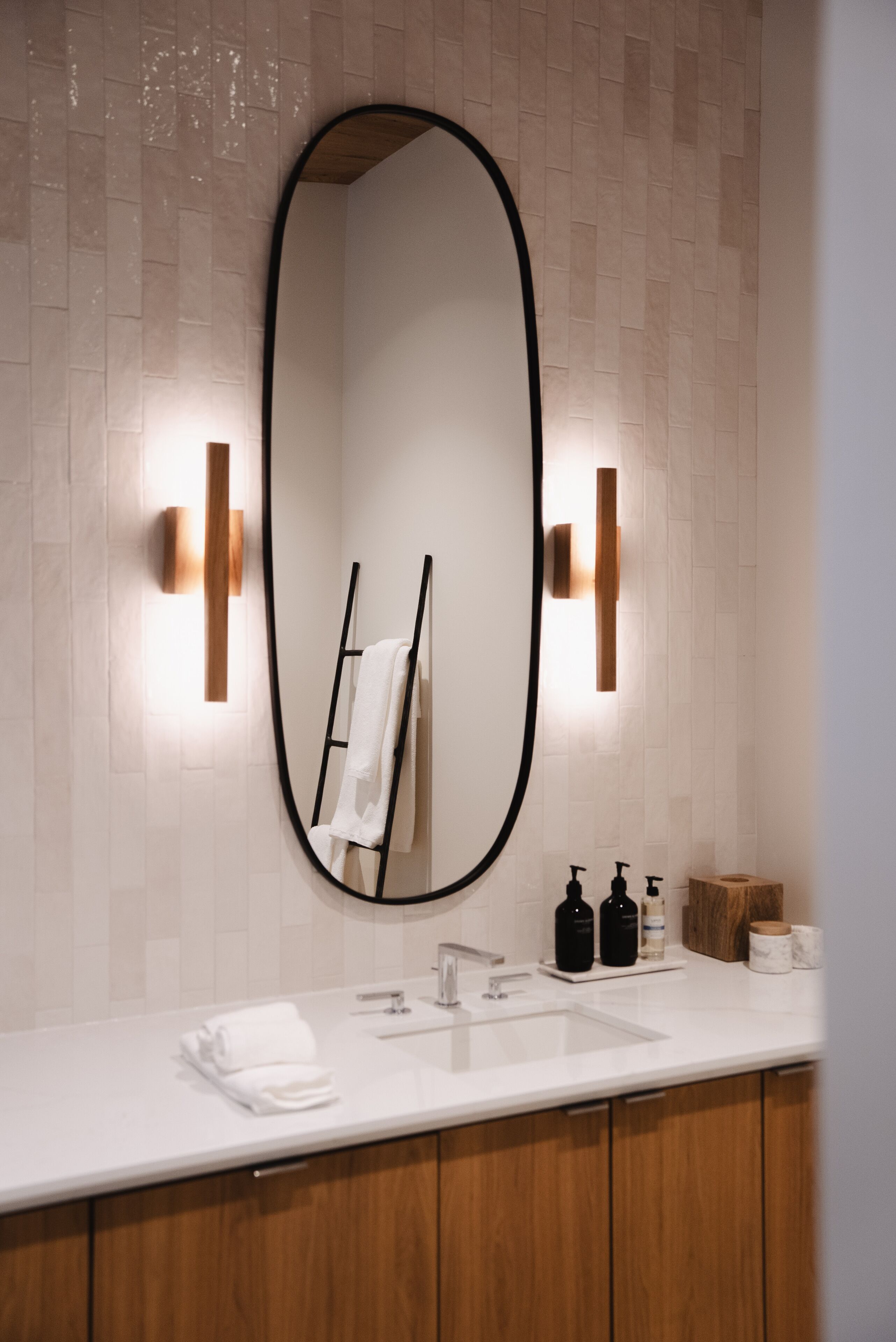 A bathroom with a large oval mirror, a white sink, wooden cabinets, and two wall-mounted soap dispensers.