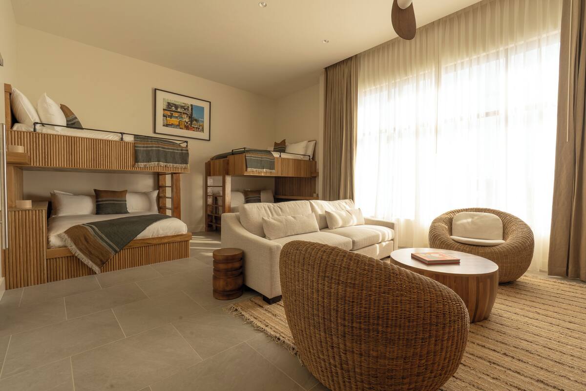 Une chambre d’hôtel moderne équipée d’un lit superposé, d’un canapé, de deux fauteuils en osier, d’une table basse et d’une grande fenêtre avec des rideaux.