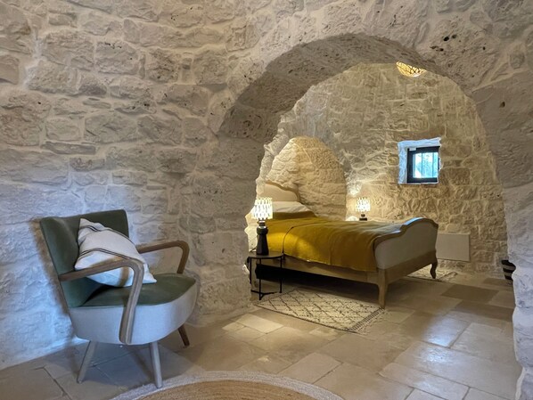 A room with a stone wall, a bed with a yellow blanket, a chair, and a small window.