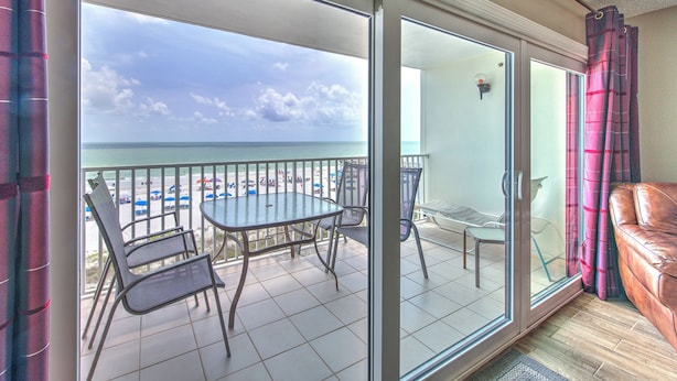 Ein Balkon mit Glastür, Tisch und Stühlen mit Blick auf einen Strand mit Sonnenschirmen und einem klaren Himmel.