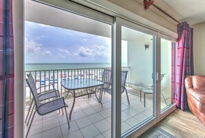 A balcony with a glass door, a table, and chairs, overlooking a beach with umbrellas and a clear sky. - Beach Palms 306 is a Gold Tier Gulf Front Condo (Indian Shores)