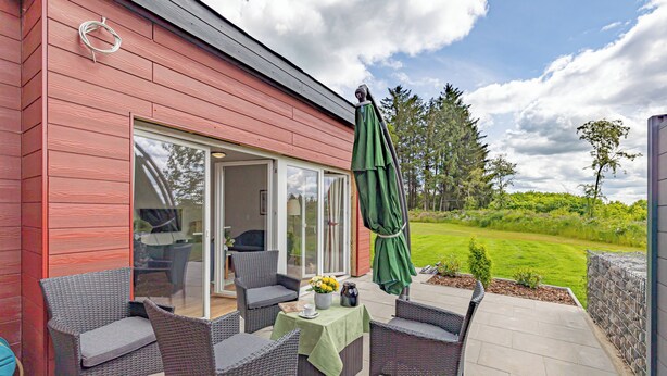 An outdoor patio with wicker furniture and a green umbrella, a red building wall, and a view of a grassy area.