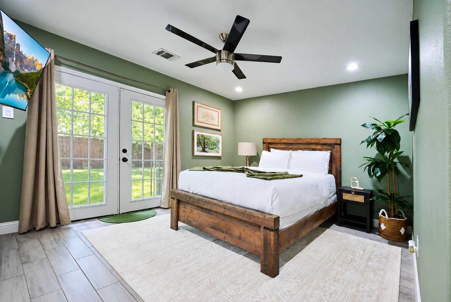 A bedroom with a bed, a ceiling fan, a nightstand, a lamp, a window with curtains, and a view of the outdoors.
