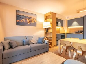 A modern living room with a sofa, a dining table with chairs, a lamp, and a kitchen area. - Wifi, 150m from resort center, swimming pool, sauna, hammam, balcony, parking, tv, ski locker, 54m² (Vaujany)
