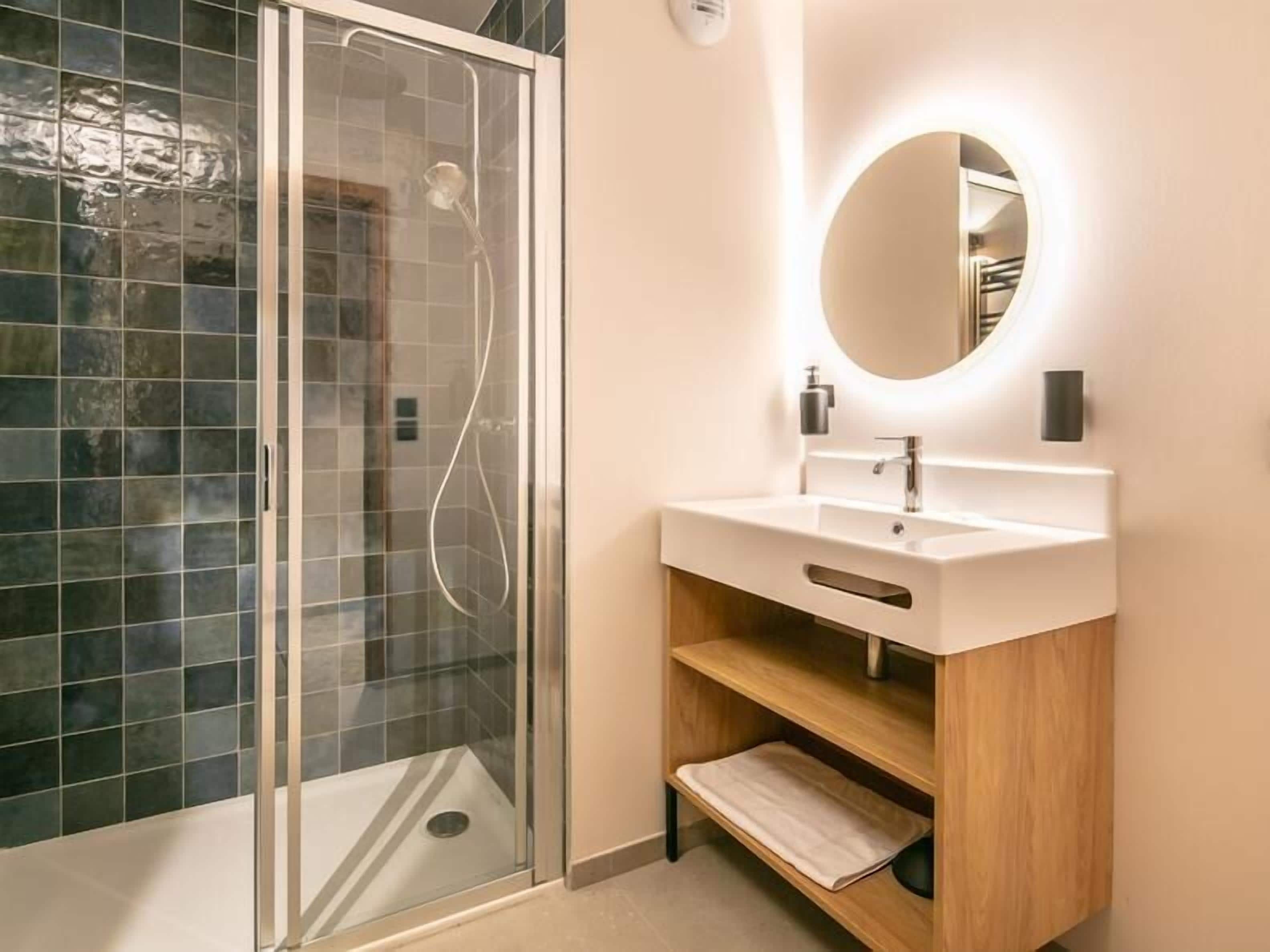 Een moderne badkamer met een douchecabine van glas, een wit wastafelmeubel met een houten onderkast en een ronde spiegel.