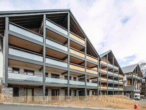 Un edificio de varios pisos con balcones, fachada de piedra y montañas al fondo.