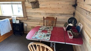 Interior de una cabaña de madera con una mesa roja, una silla, otra silla de madera, un ventanal y una estufa.