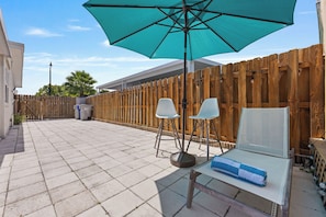 A patio with a wooden fence, two white chairs, a table with a towel, and a large umbrella.