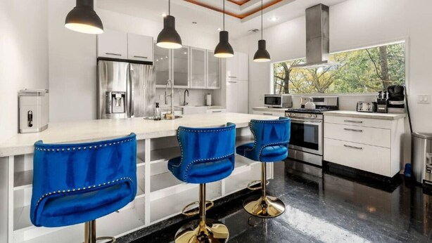 A modern kitchen with white cabinets, a stainless steel refrigerator, a gas stove, and a microwave. There are three blue bar stools with gold legs. - Modern Retreat Near Atlantic Station (Atlanta)