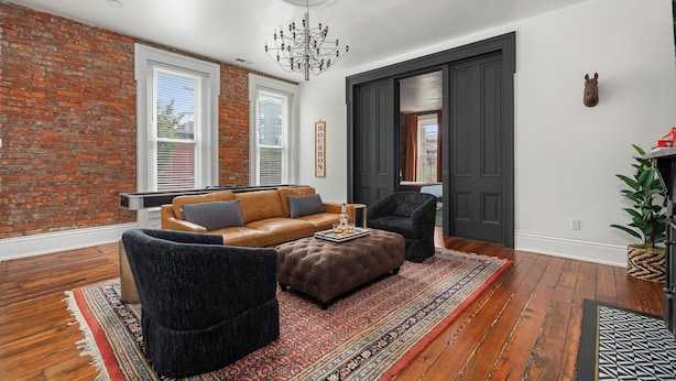 Un salon moderne avec un mur en briques, un canapé en cuir, des fauteuils foncés, un lustre et un tapis à motifs.