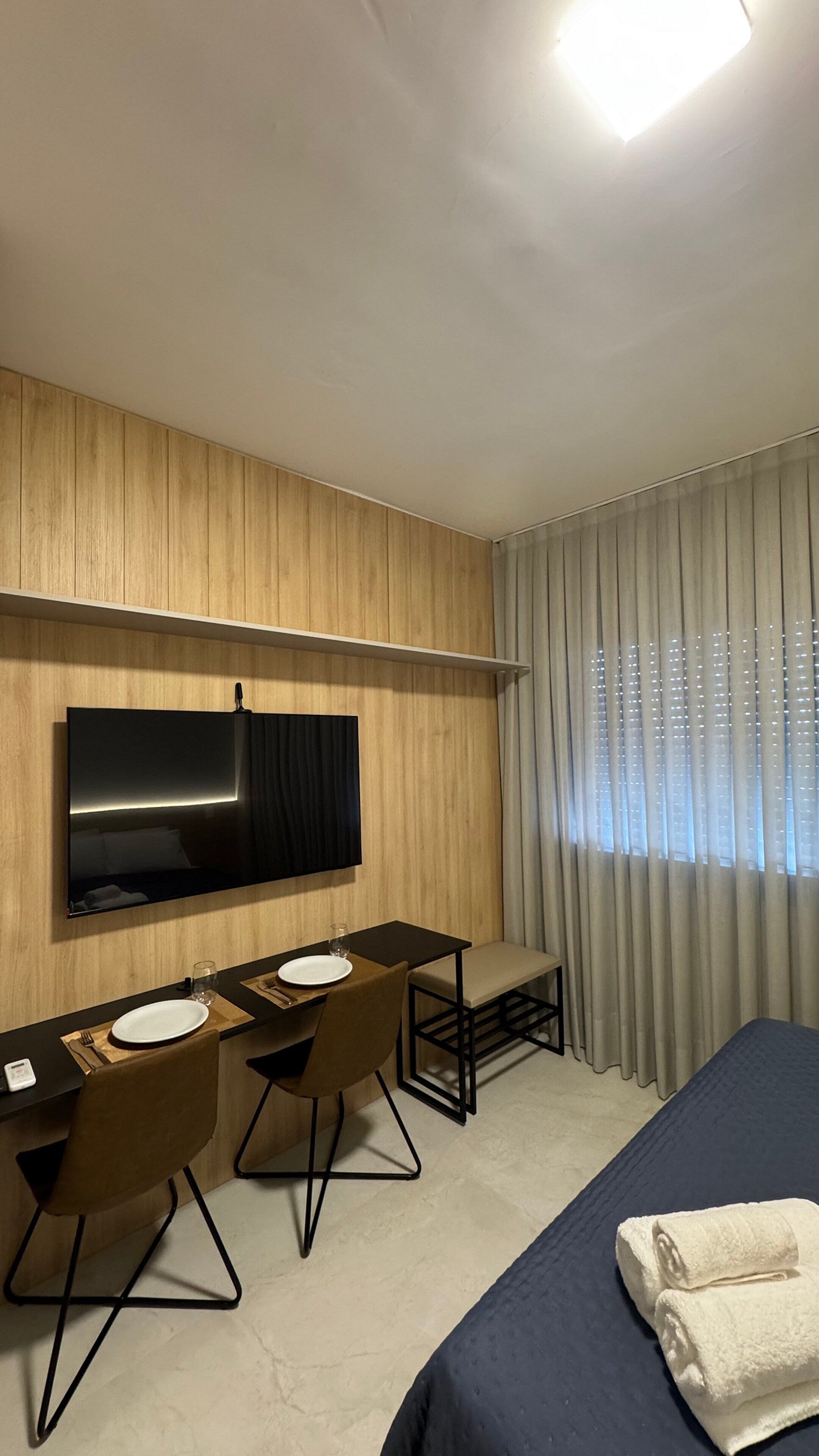 Ein Hotelzimmer mit Bett, Schreibtisch mit zwei Stühlen, Flachbildfernseher an der Wand und einem Fenster mit Vorhängen.
