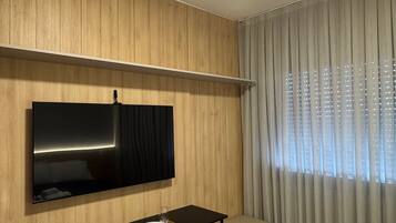 A hotel room with a bed, a desk with two chairs, a flat-screen TV mounted on the wall, and a window with curtains.