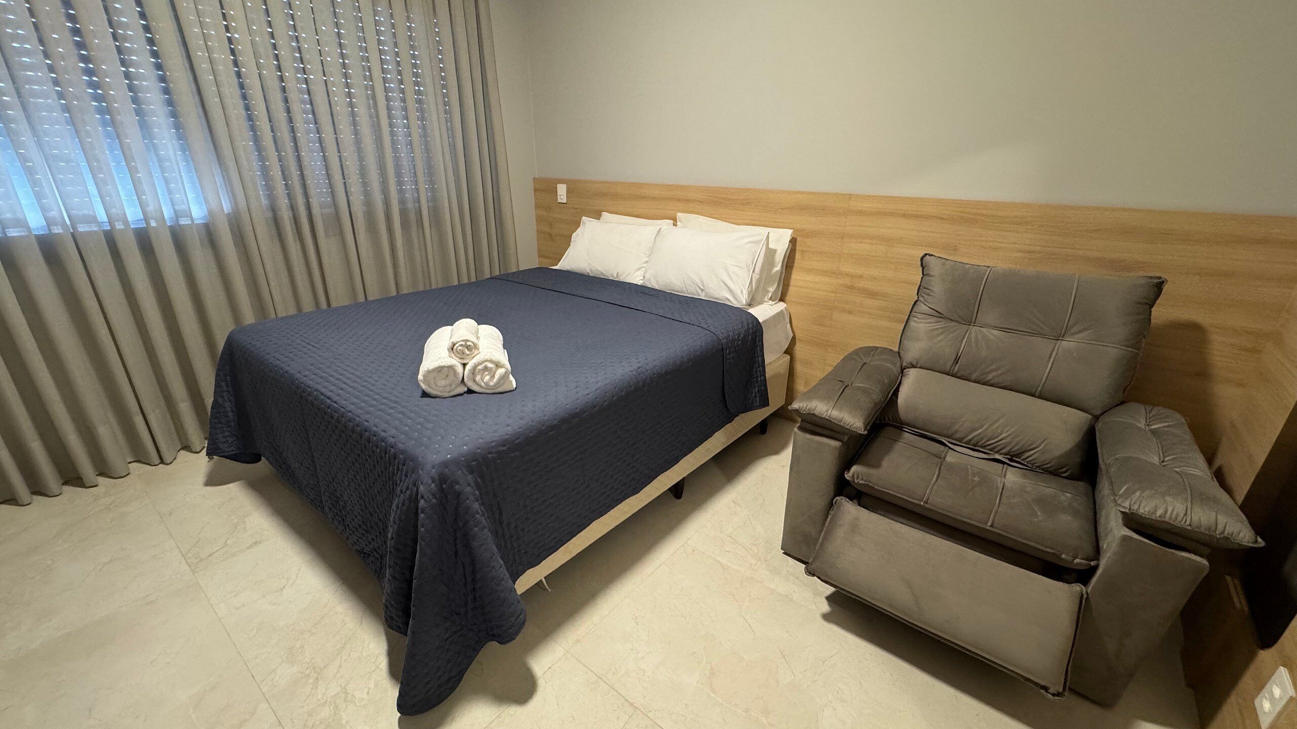 A hotel room with a bed, a massage chair, and a window with blinds.