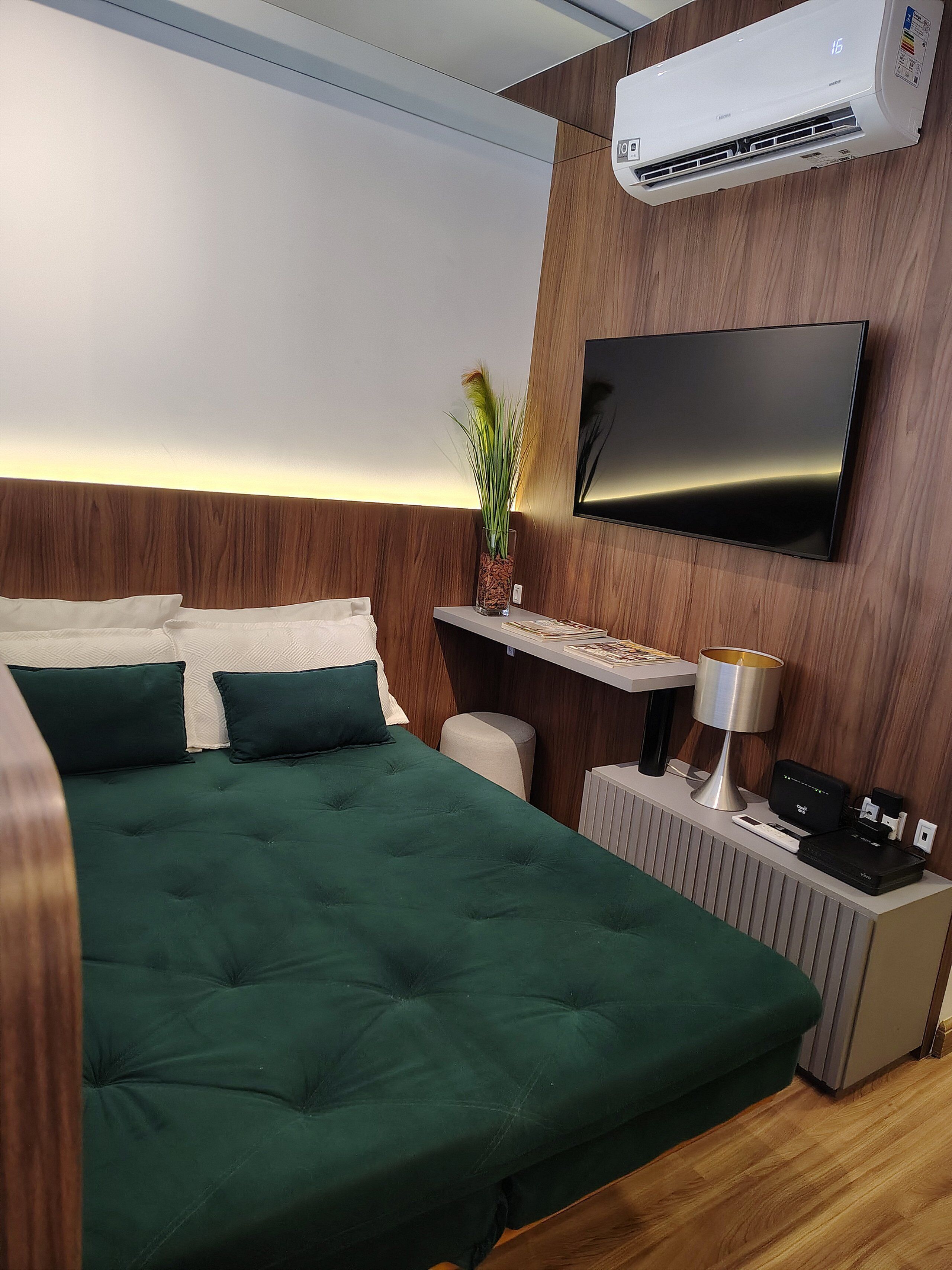 Quarto de hotel moderno com uma cama grande, televisão montada, aparelho de ar condicionado, mesa de cabeceira com luminária e uma pequena planta.