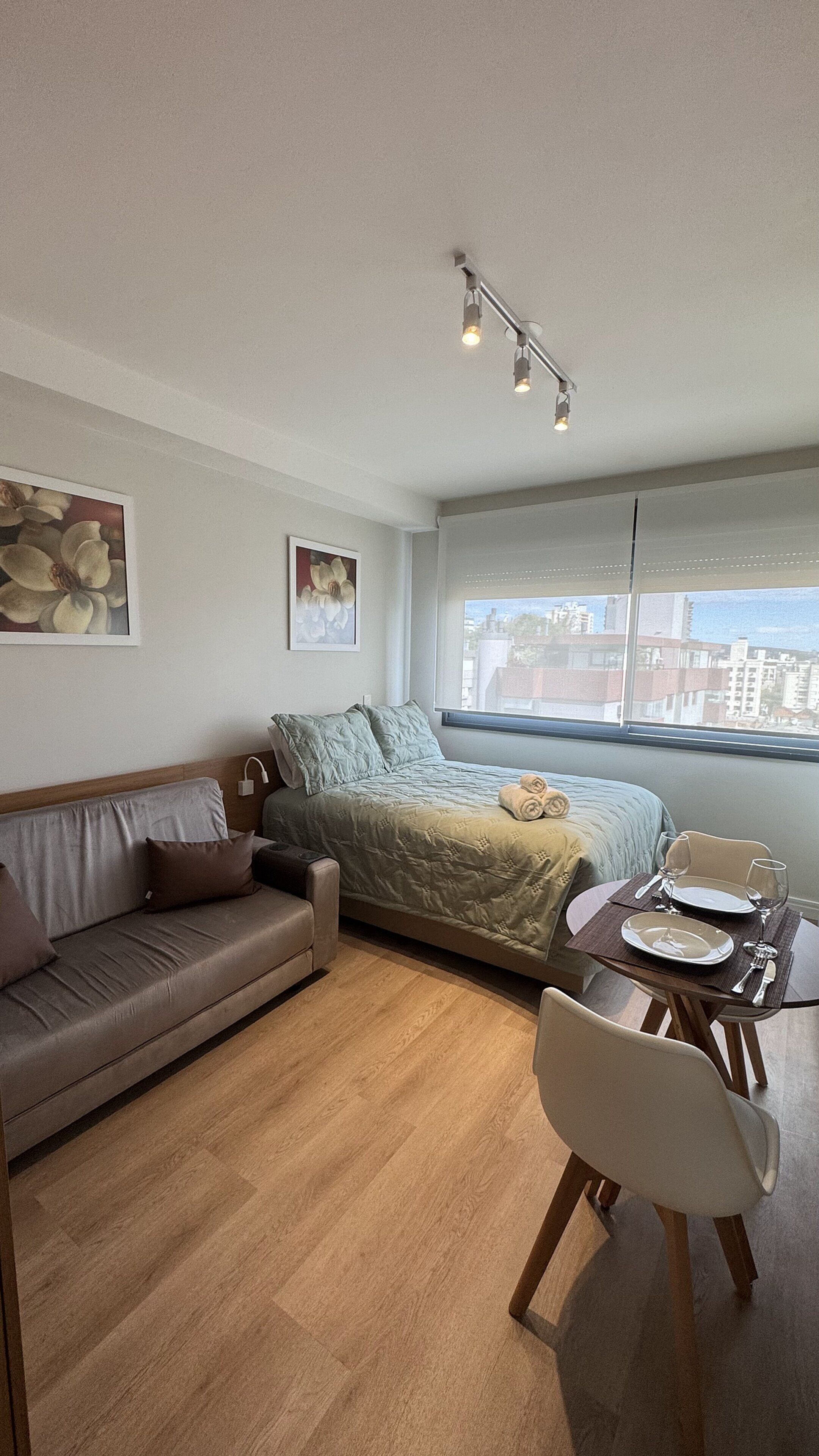 Quarto moderno com cama, sofá, mesa de jantar com cadeiras e vista para a cidade pela janela.