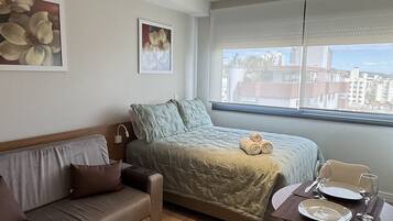 Quarto moderno com cama, sofá, mesa de jantar com cadeiras e vista para a cidade pela janela.