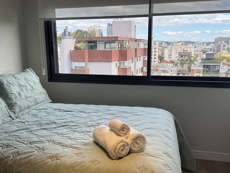 A bedroom with a large window, a bed with a quilt, and a rolled towel on the bed.