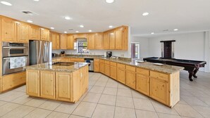 A spacious kitchen with wooden cabinets, granite countertops, stainless steel appliances, and a pool table.