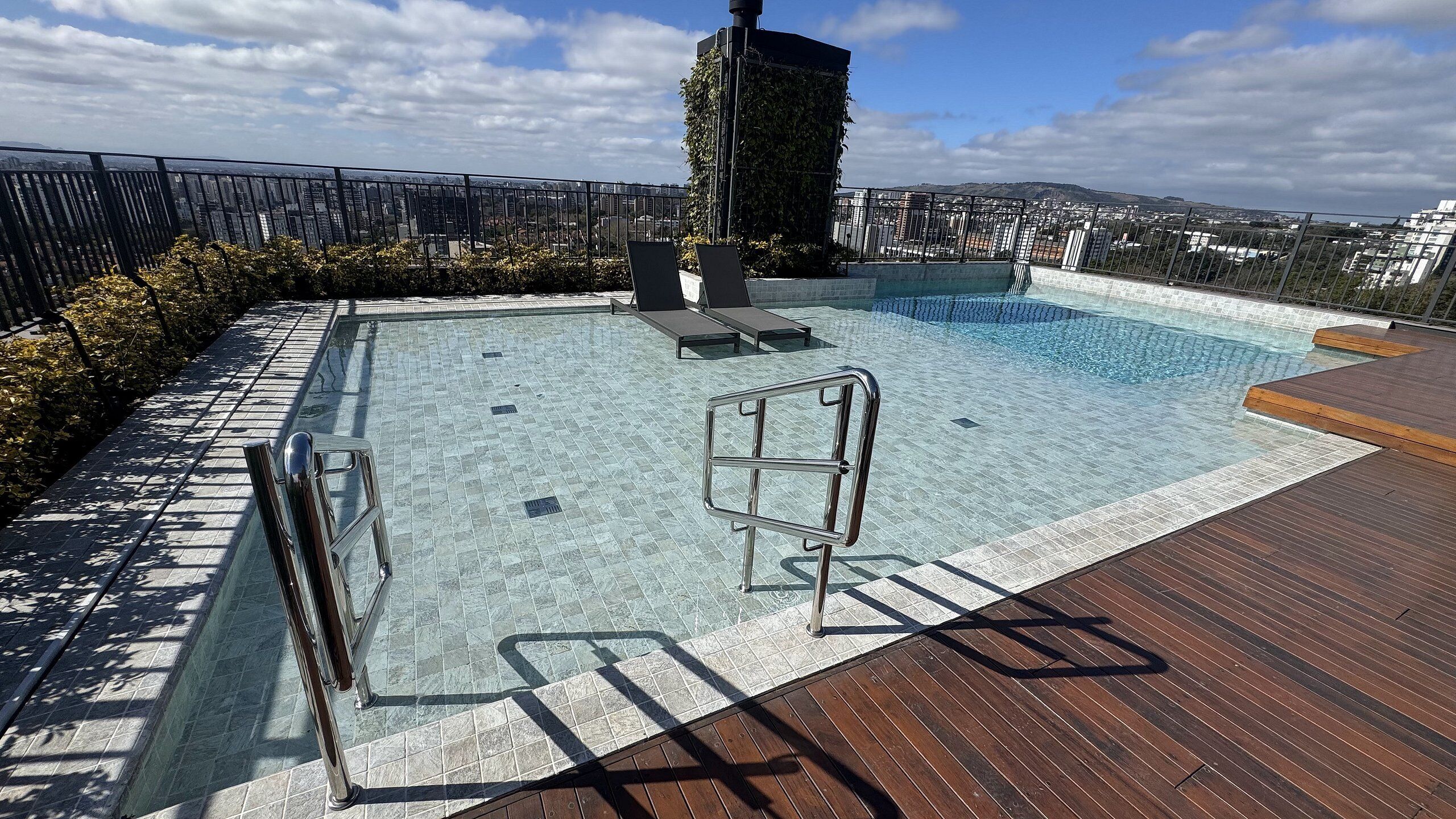 A rooftop swimming pool with a wooden deck, a metal safety rail, and a view of the cityscape.