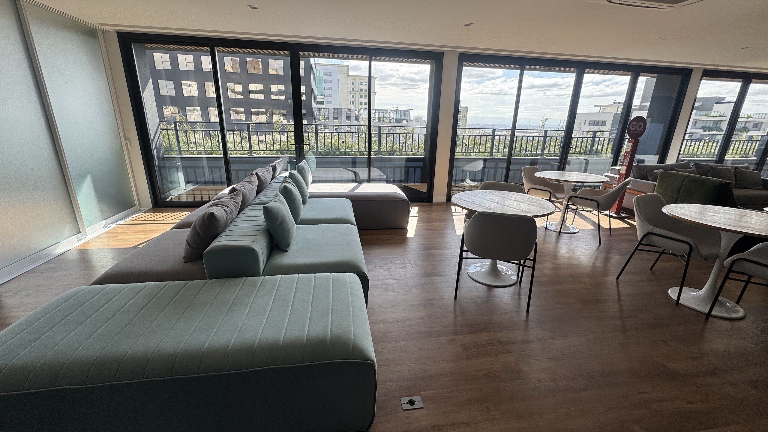 A modern lounge area with a long sofa, multiple chairs, and a small table. Large windows offer a view of the cityscape.