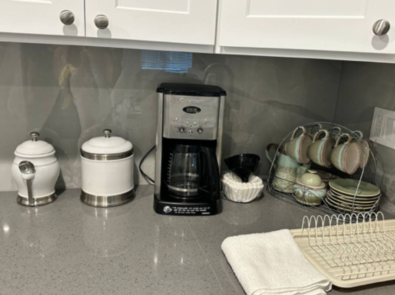 A kitchen with a coffee maker, two containers, a dish rack, and a towel.