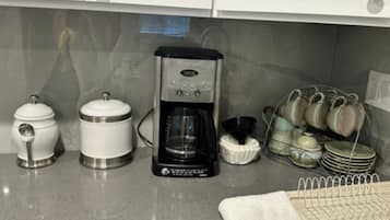 A kitchen with a coffee maker, two containers, a dish rack, and a towel.