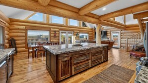 A spacious log cabin interior with a large kitchen island, wooden floors, and a view through the windows. - Here & Now | Luxe Yellowstone Cabin w/ Hot Tub & Views! (Island Park)