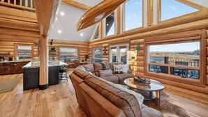 A log cabin interior with a large living area, a kitchen, and large windows offering views of the landscape. - Here & Now | Luxe Yellowstone Cabin w/ Hot Tub & Views! (Island Park)
