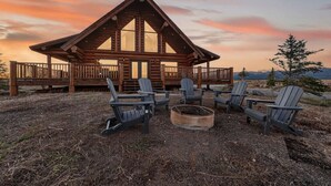 A log cabin with a deck, surrounded by Adirondack chairs and a fire pit, set against a sunset backdrop. - Here & Now | Luxe Yellowstone Cabin w/ Hot Tub & Views! (Island Park)