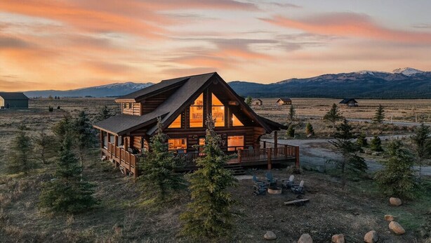 A log cabin with a deck, surrounded by trees and mountains in the background. - Here & Now | Luxe Yellowstone Cabin w/ Hot Tub & Views! (Island Park)