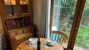 Un coin repas avec une table ronde en bois, dressée pour quatre personnes, entourée de chaises à motifs floraux. Un buffet en bois, doté de portes en verre et contenant des livres, se trouve à gauche.
