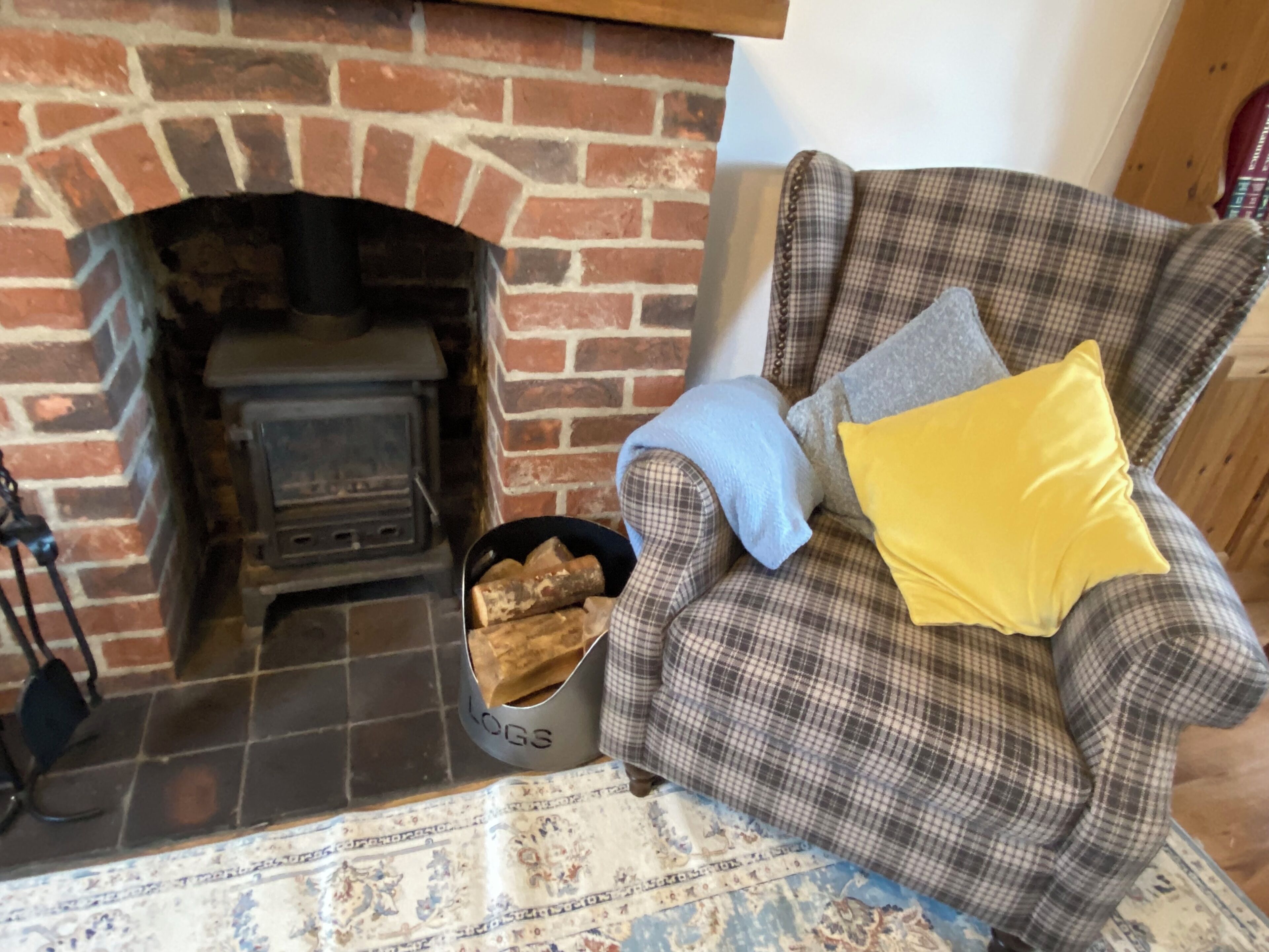 A cozy living room with a brick fireplace, a plaid armchair with a yellow pillow, and a basket of logs.