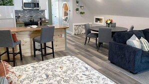 Une cuisine moderne dotée d’un îlot central, de tabourets de bar, d’un coin repas avec une table ronde et des chaises, ainsi que d’un canapé bleu foncé agrémenté de coussins décoratifs à motifs floraux.
