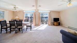 A spacious living room with a dining area, large windows, ceiling fans, and a television. - Catskills Lakefront Getaway at Sackett Lake (Monticello)