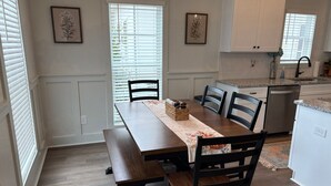 Un coin repas comprenant une table en bois, quatre chaises et un banc. Il y a deux fenêtres équipées de stores, une cuisine avec des meubles blancs et un plan de travail avec un évier.