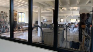 A modern interior with a large glass window, a seating area with a sofa and chairs, a dining area with a table and chairs, and a visible kitchen area.