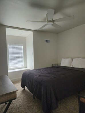 A bedroom with a bed, a ceiling fan, a window with blinds, a bedside table, and a carpeted floor.