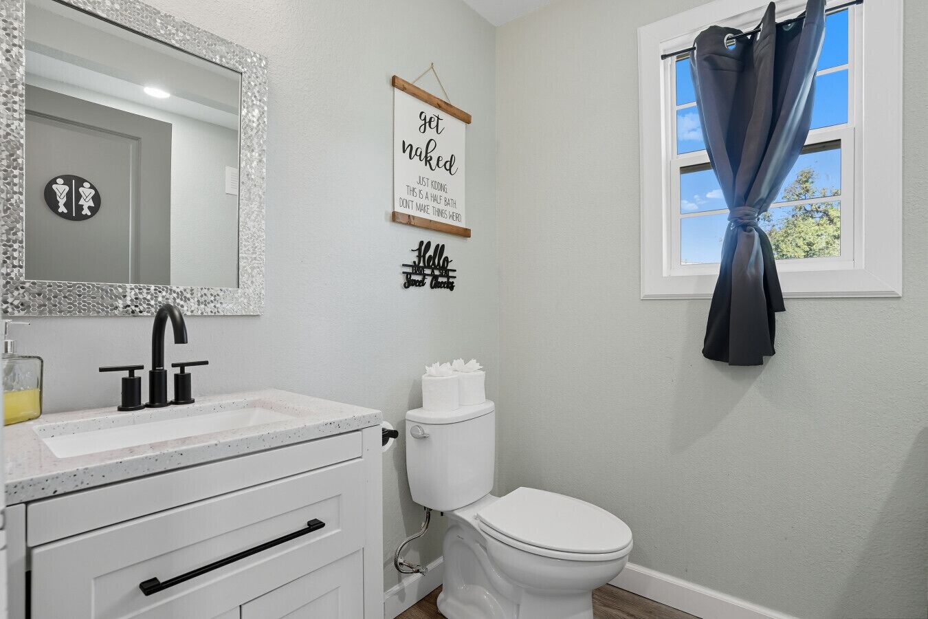 A bathroom with a white sink, black faucet, and a toilet. There is a sign on the wall that says 'get naked' and another sign that says 'Hello, Sweetie.'