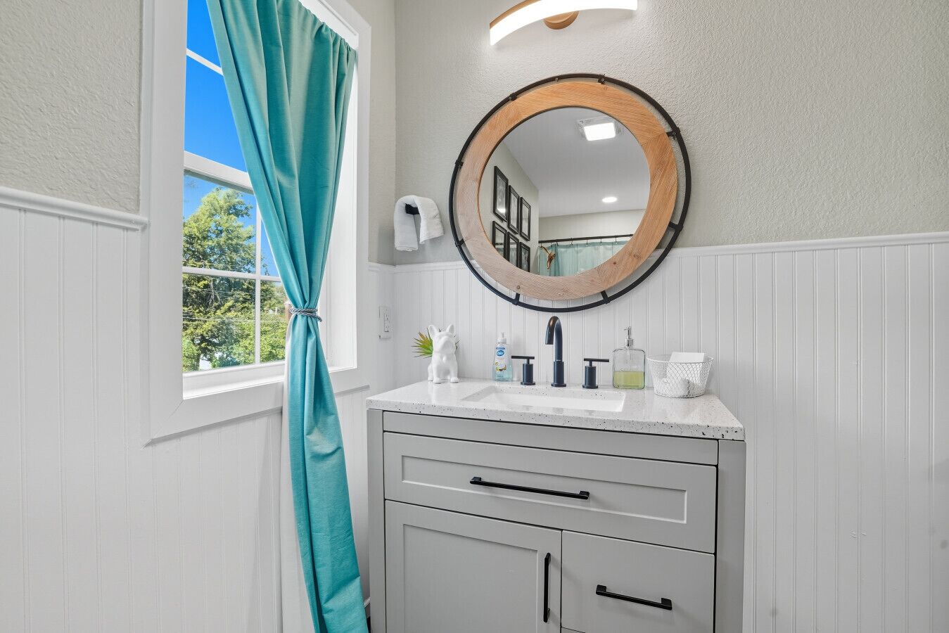 Une salle de bain avec un meuble-lavabo blanc, un miroir rond, une fenêtre ornée de rideaux turquoise et une vue sur la verdure à l’extérieur.