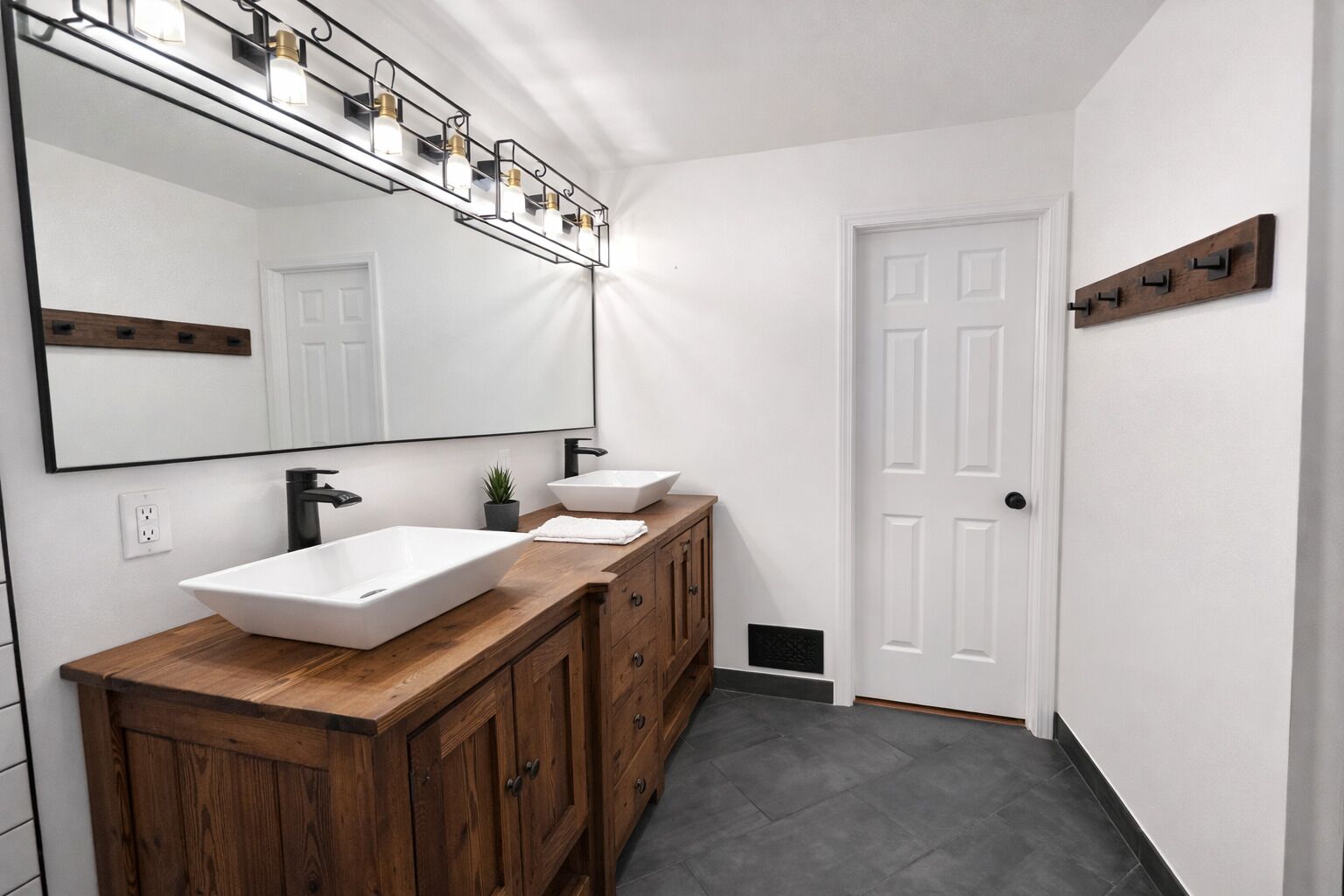 A bathroom with a large mirror, two sinks, wooden cabinets, a potted plant, and a white door.