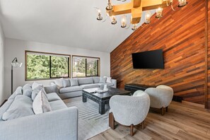 A modern living room with a sectional sofa, a coffee table, and two armchairs. There is a flat-screen TV mounted on a wooden wall, and a floor lamp is visible.