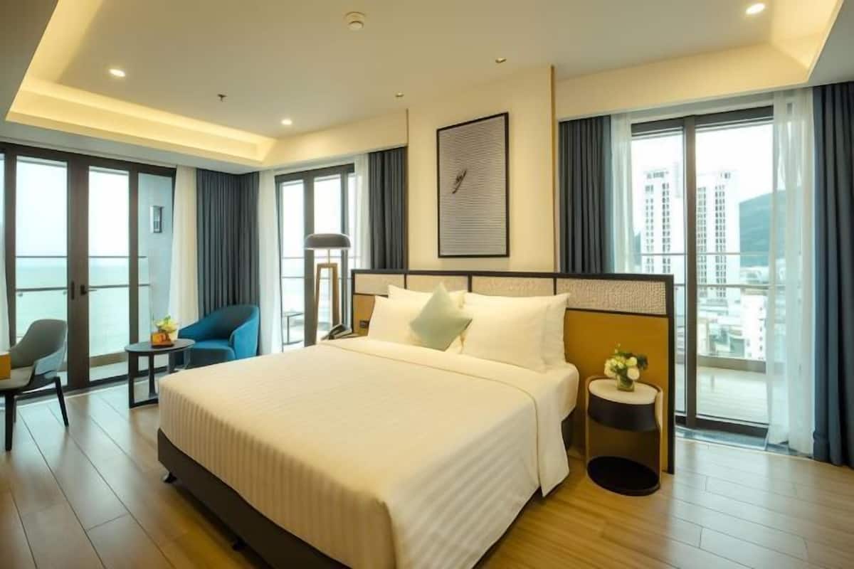 A hotel room with a large bed, a desk, a chair, a small table with flowers, and a view of buildings through large windows.