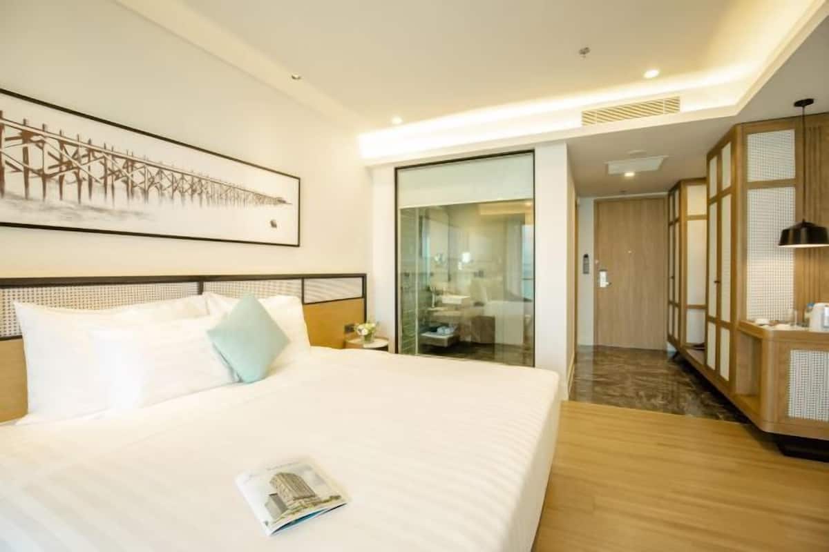 A hotel room with a large bed, a bedside table, a nightstand, a framed artwork on the wall, a sliding glass door leading to a bathroom, and a wooden cabinet.