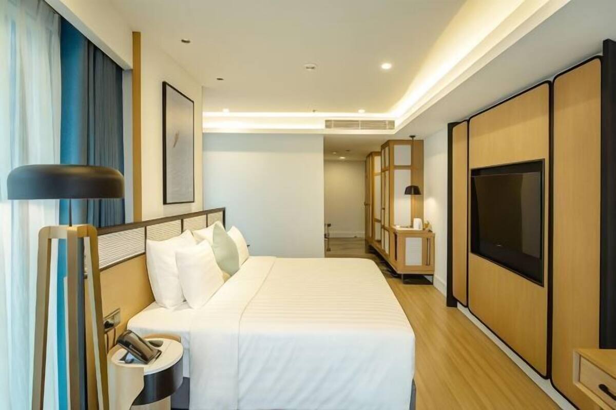 A hotel room with a bed, bedside table, lamp, TV, and a view of the hallway.