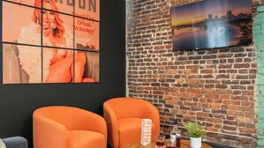 A room with two orange armchairs, a wooden coffee table with drinks and a plant, a brick wall, and a cityscape poster. - Stable #5 ~ Mini Golf, Fire Pits, Games, Walkable (Louisville)